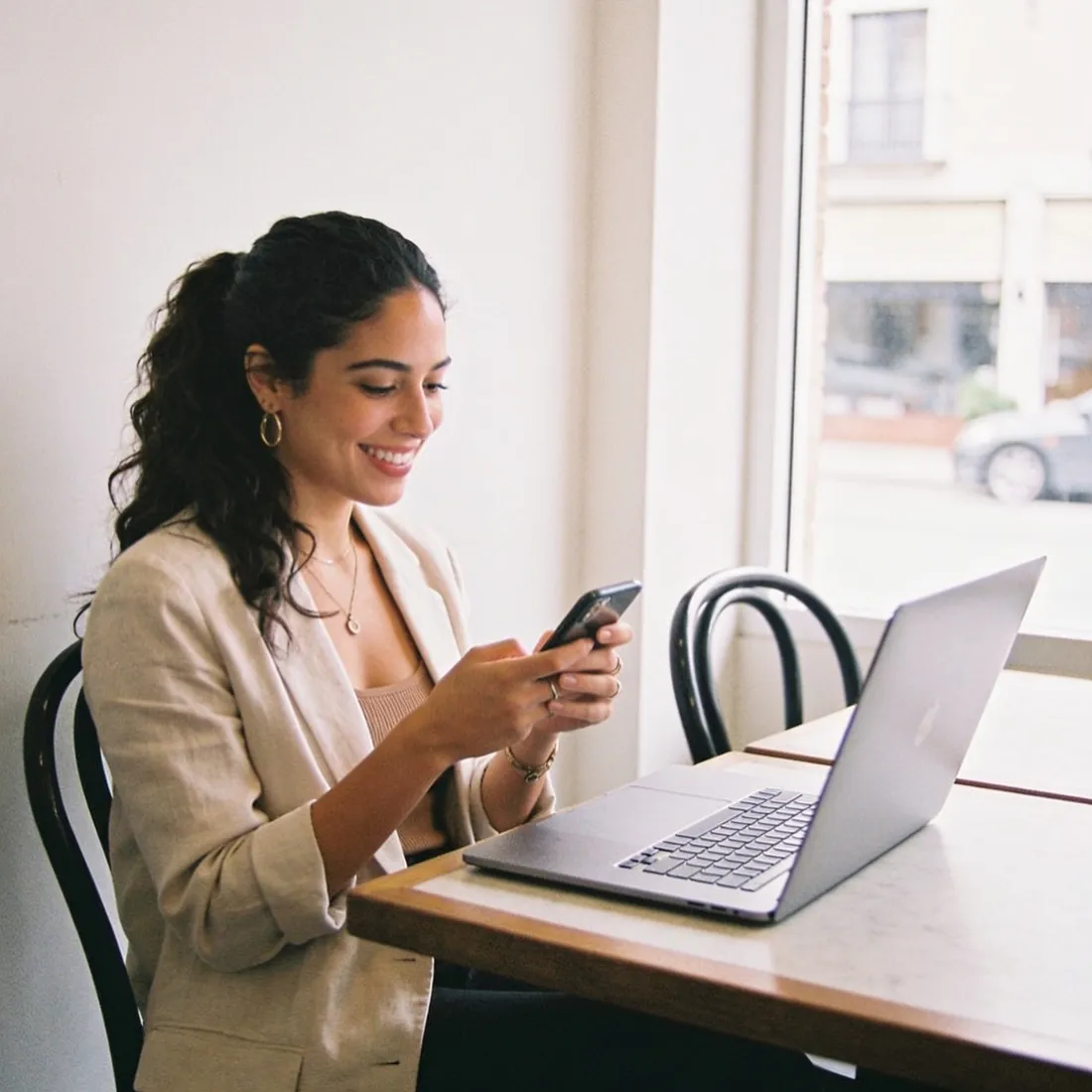 A small-business owner writing social posts on her laptop and phone in a bright cafe